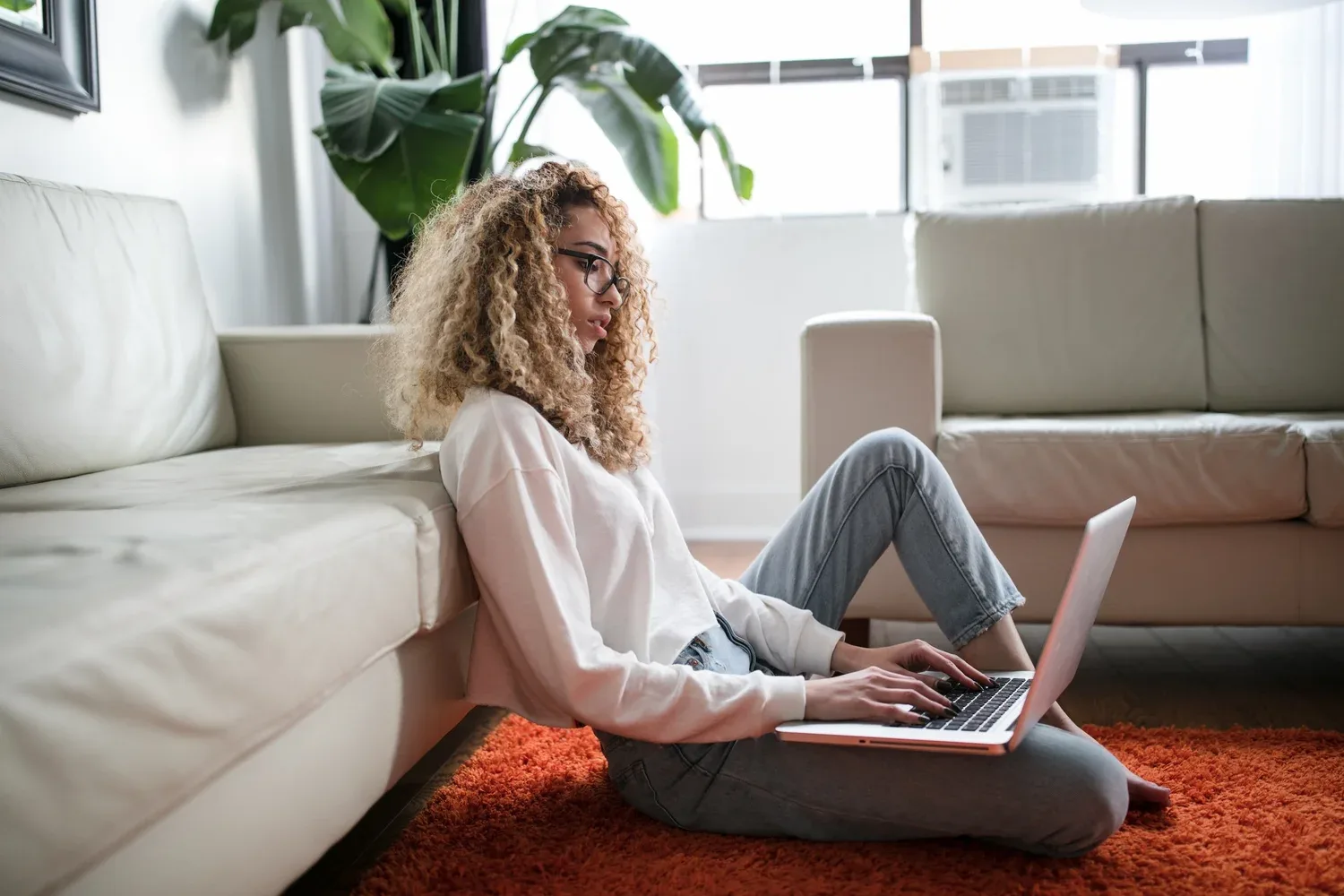 Woman sitting on the floor by a sofa working on a laptop