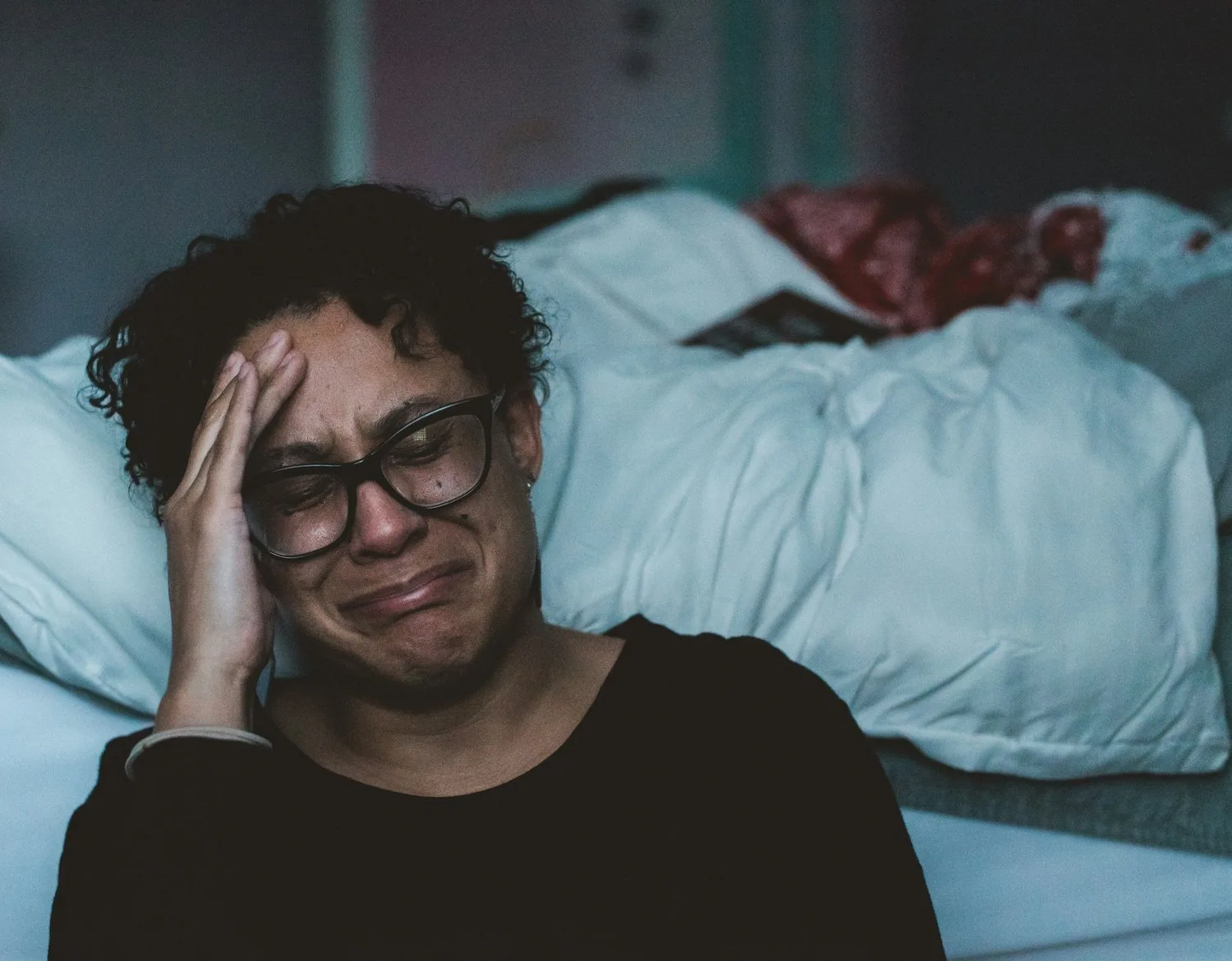Woman in glasses sitting on the floor by a bed with a hand to her head, crying