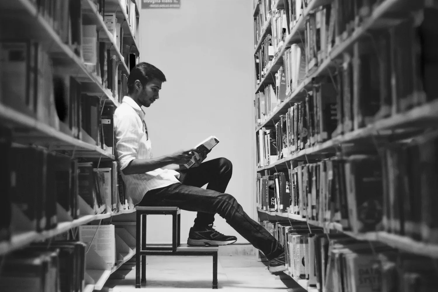 Black-and-white photo of a person reading on a bench between library shelves