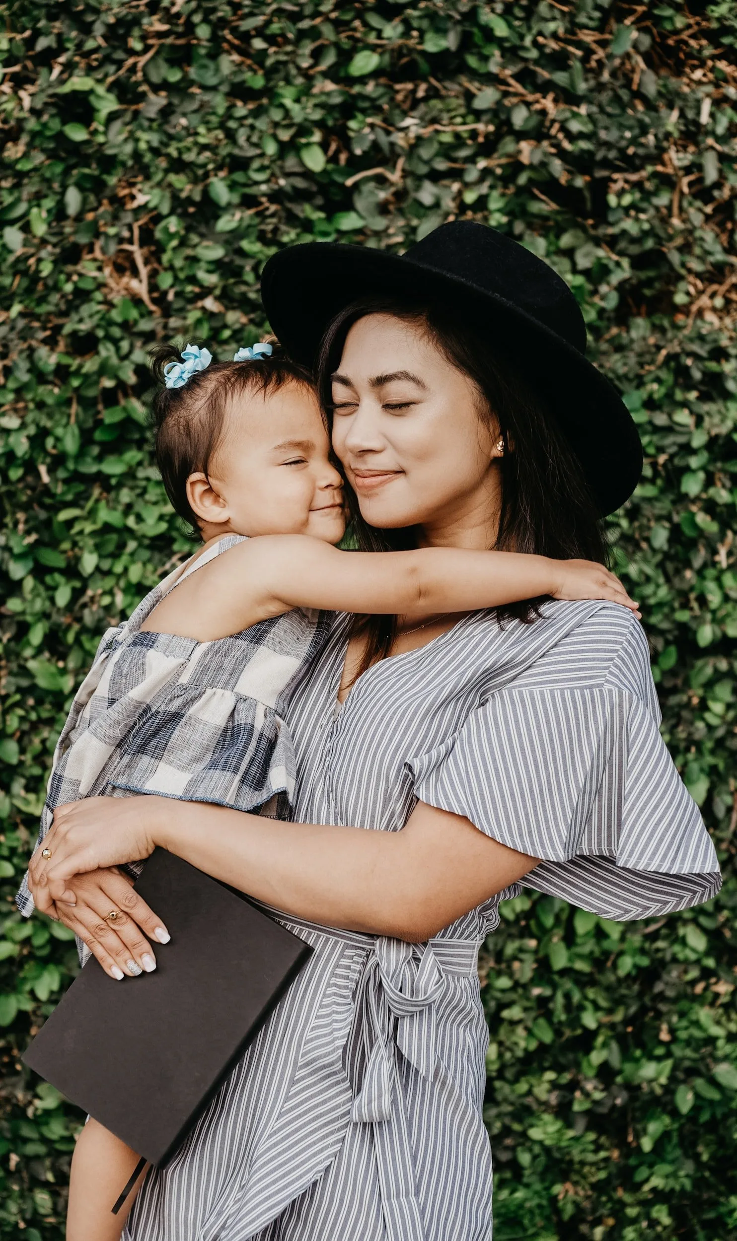 Smiling mother in a wide-brimmed hat holding her baby in front of greenery