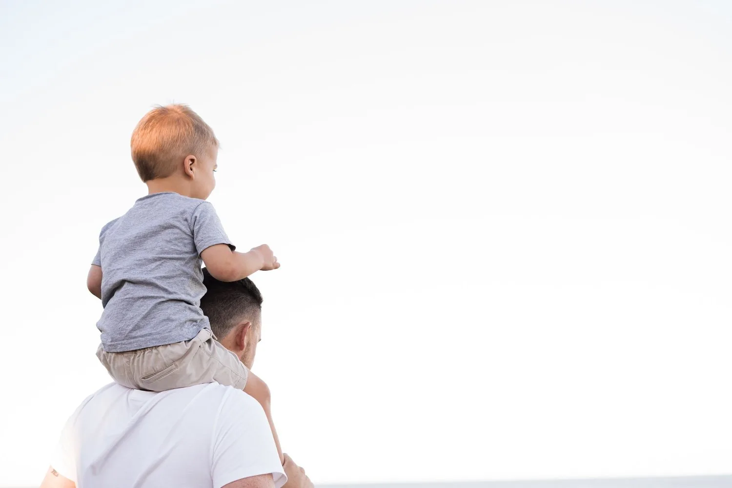 Toddler sitting on a parent’s shoulders against a bright sky