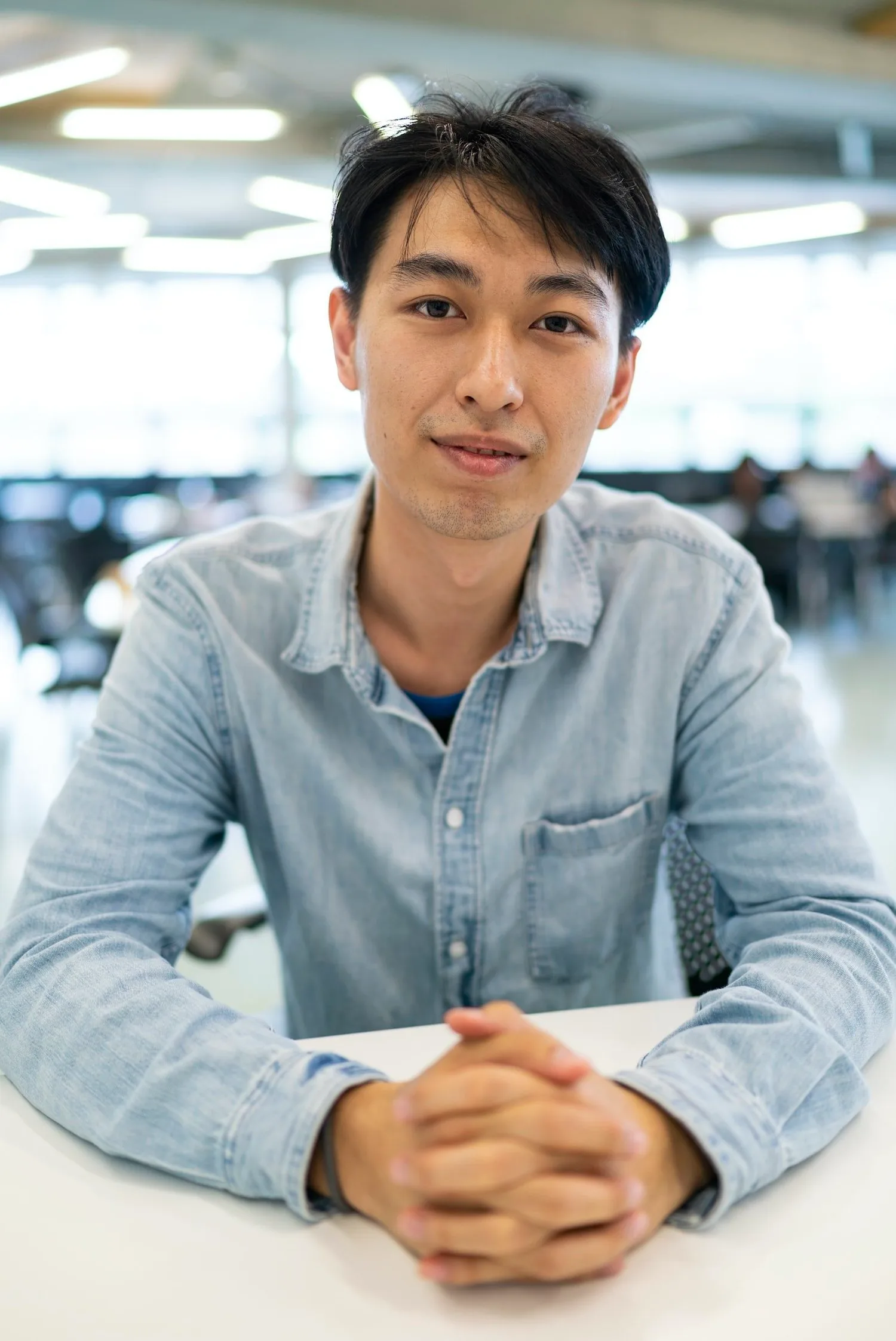Young East Asian man in a denim shirt seated at a table with hands folded