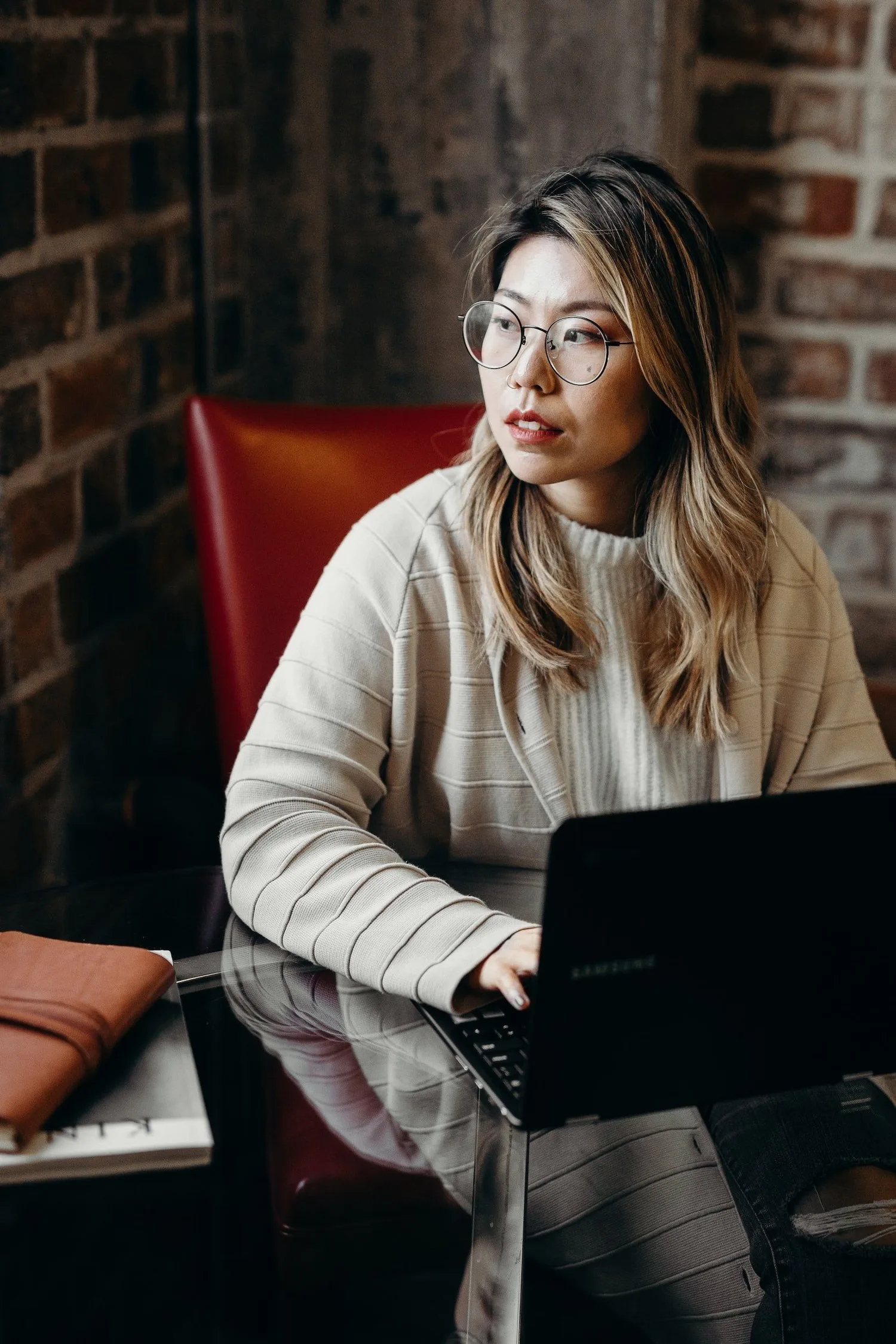 East Asian woman in glasses working on a laptop, looking away thoughtfully