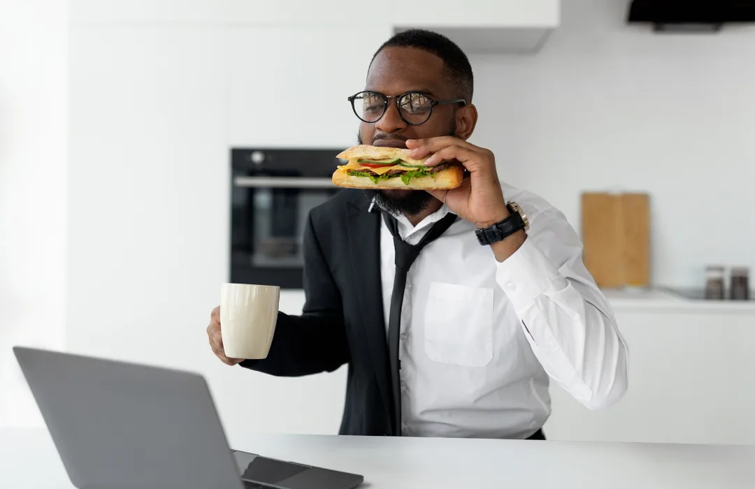 Man in a suit eating a sandwich and holding a mug at his work desk
