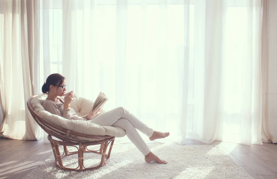 Woman reading in a rattan chair beside tall, sunlit curtains