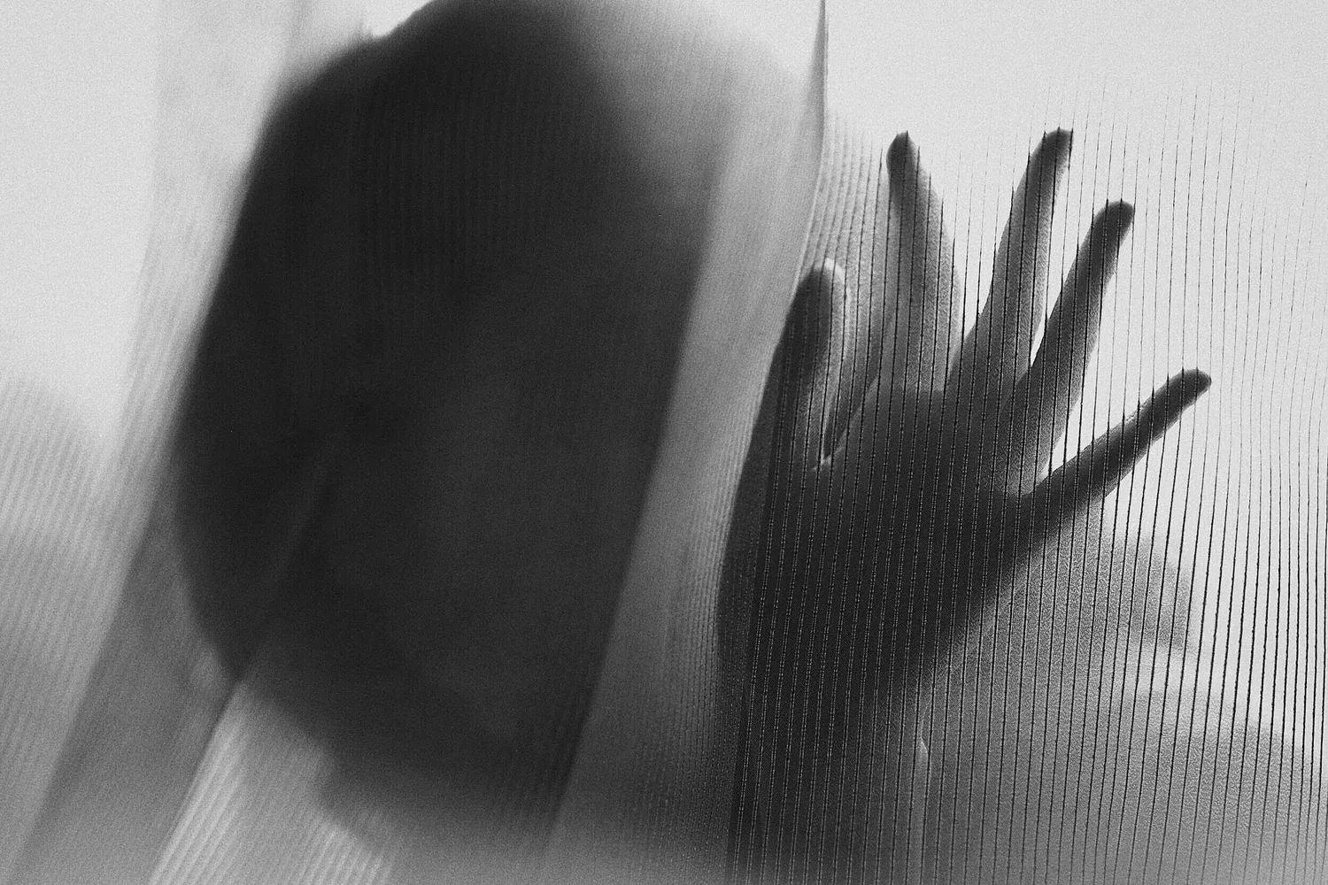 Black-and-white photo of a hand pressed against textured glass beside a blurred face