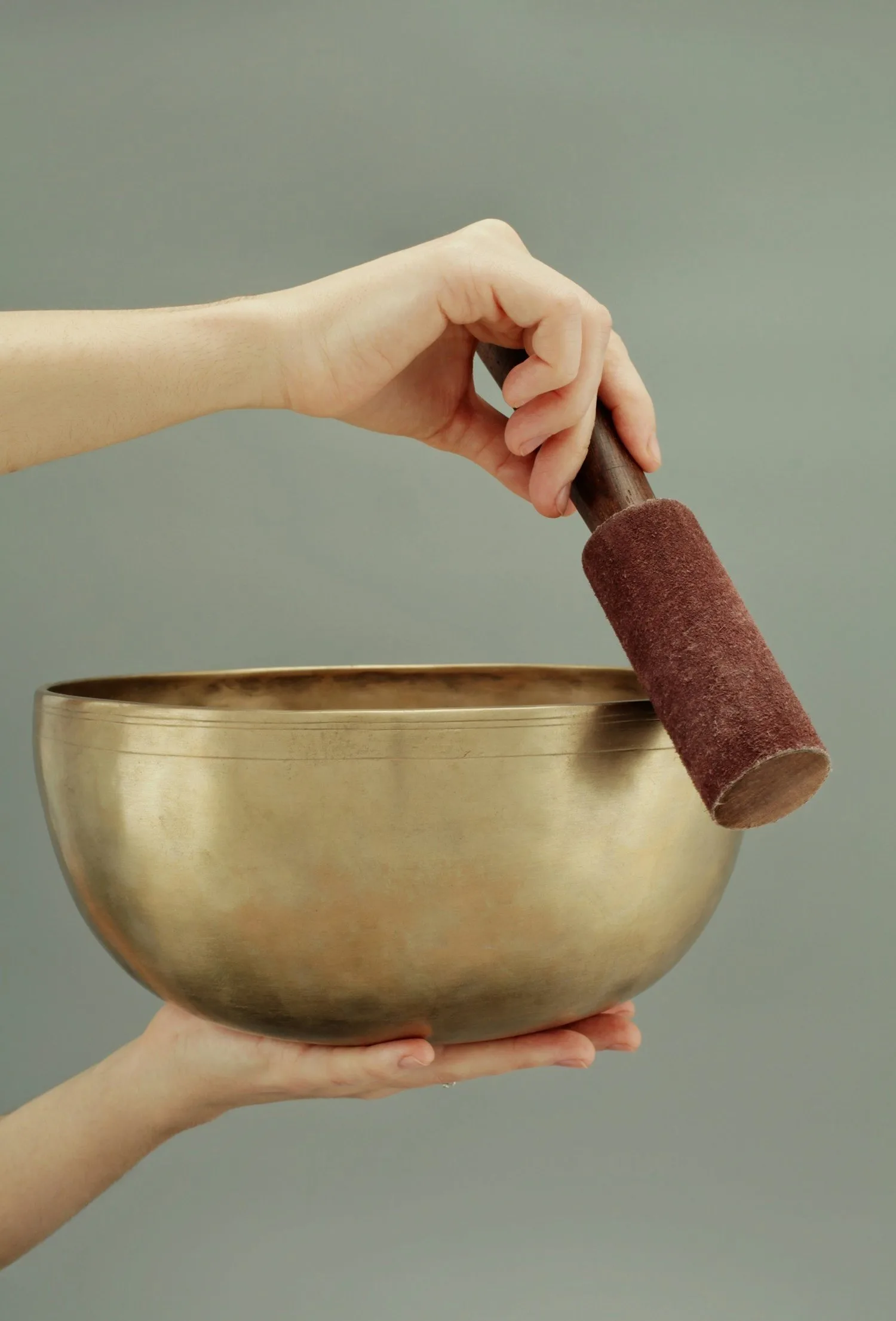 Hand holding a brass singing bowl and wooden mallet against a plain background