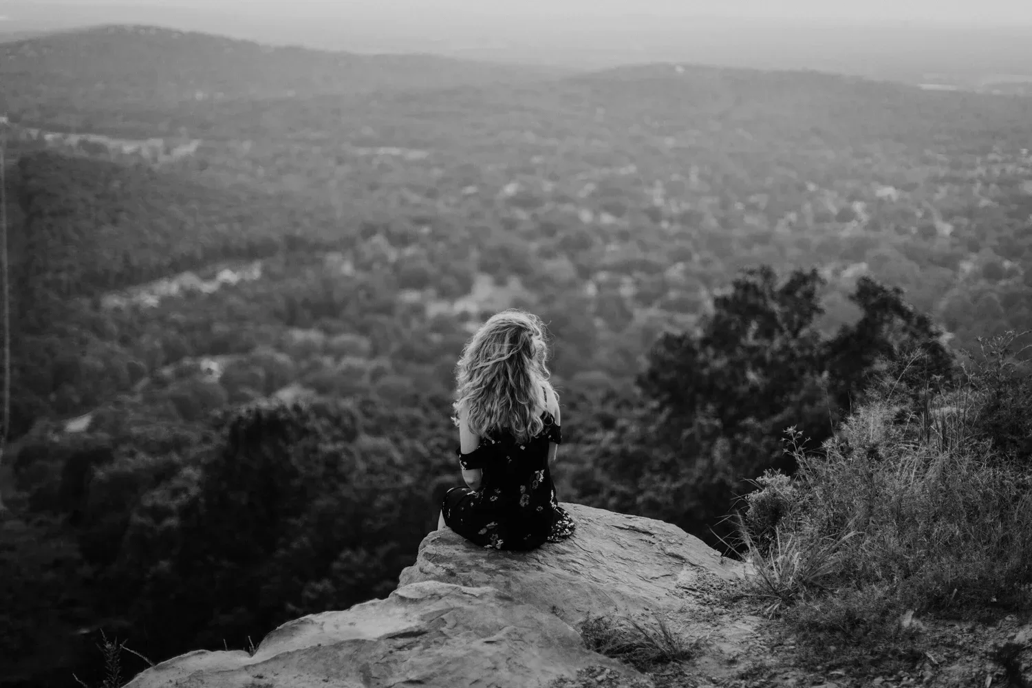Black-and-white photo of a person sitting on a rock overlooking a forested valley