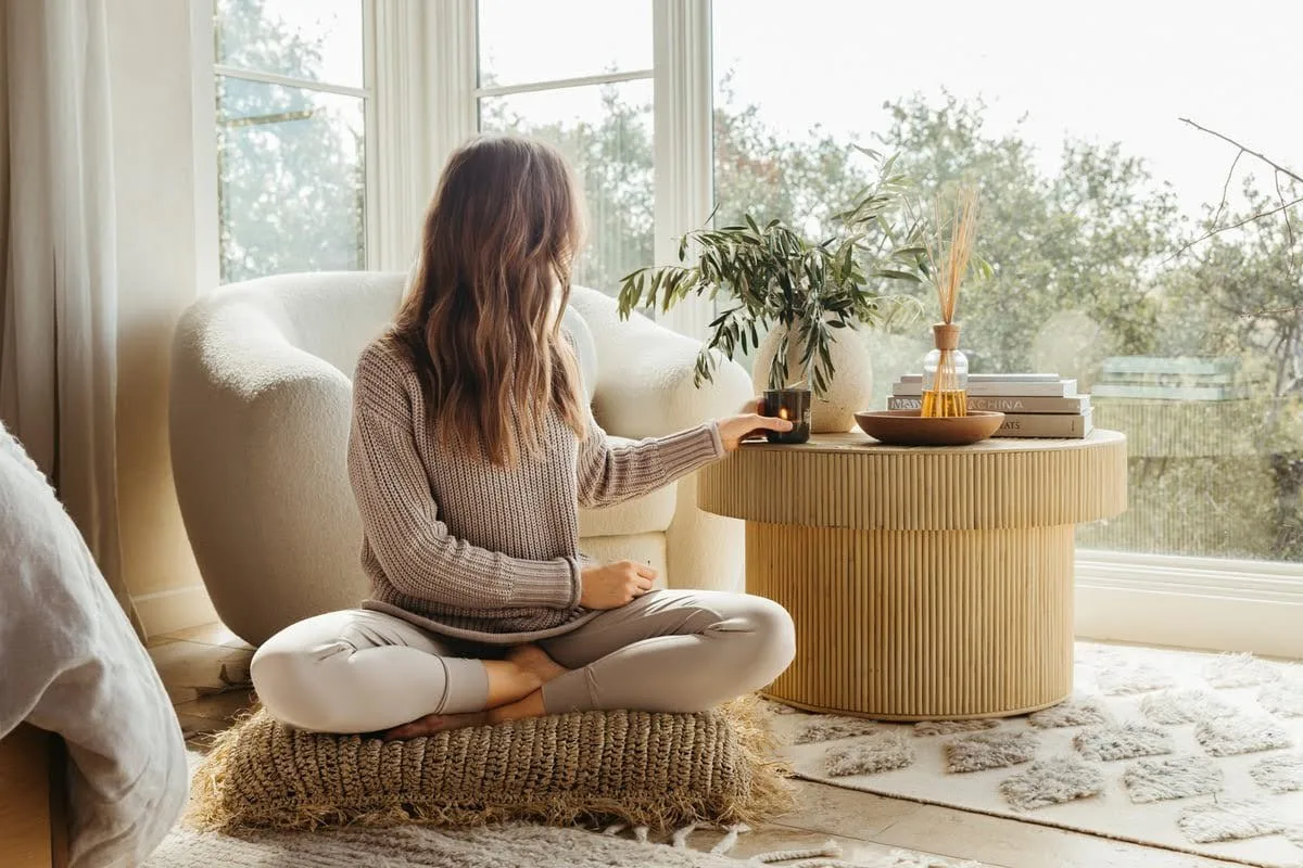 Woman sitting cross-legged on a floor cushion by a window, journaling