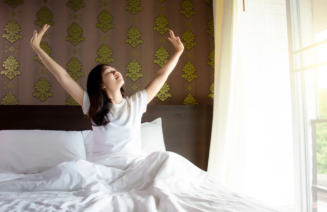 Woman stretching her arms overhead in bed by a sunny window
