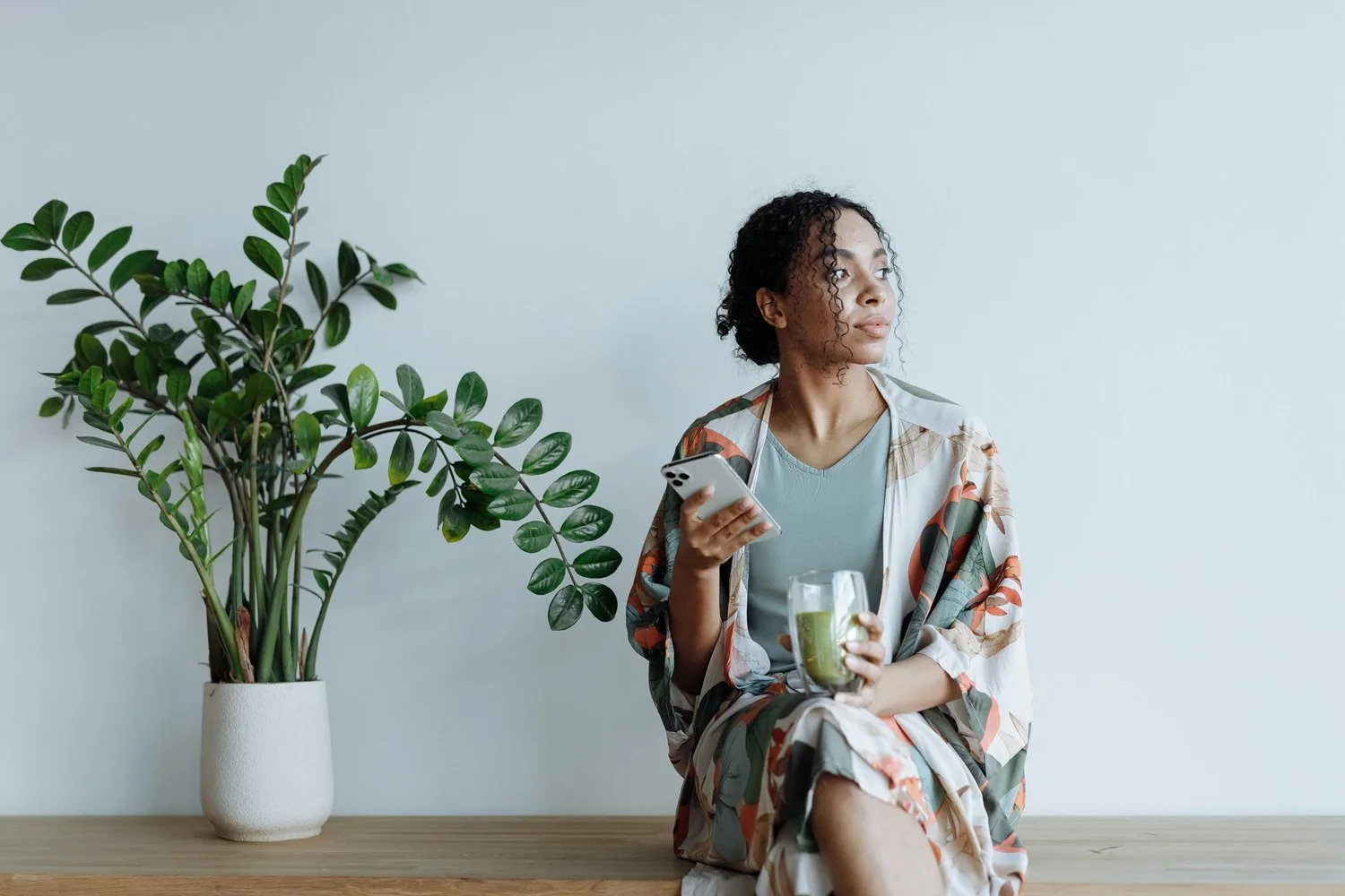 Woman in a patterned robe sitting on a bench beside a potted plant, holding a mug