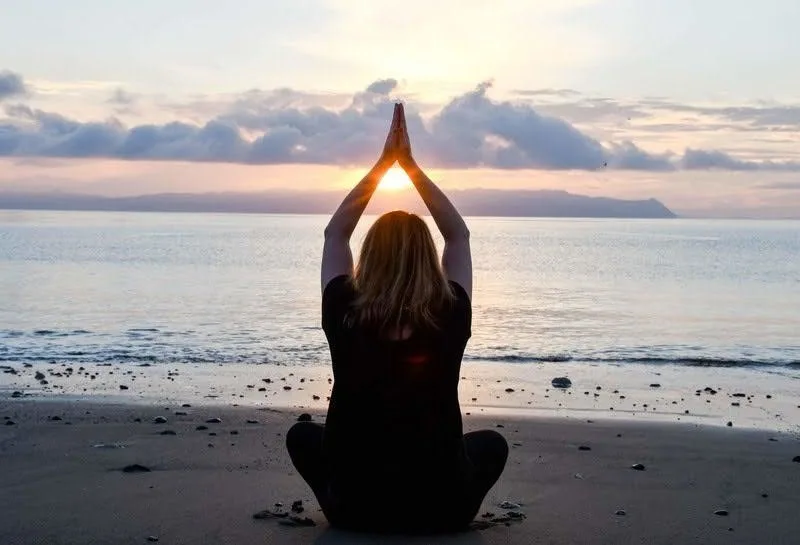 Person meditating on a beach at sunset with hands raised overhead
