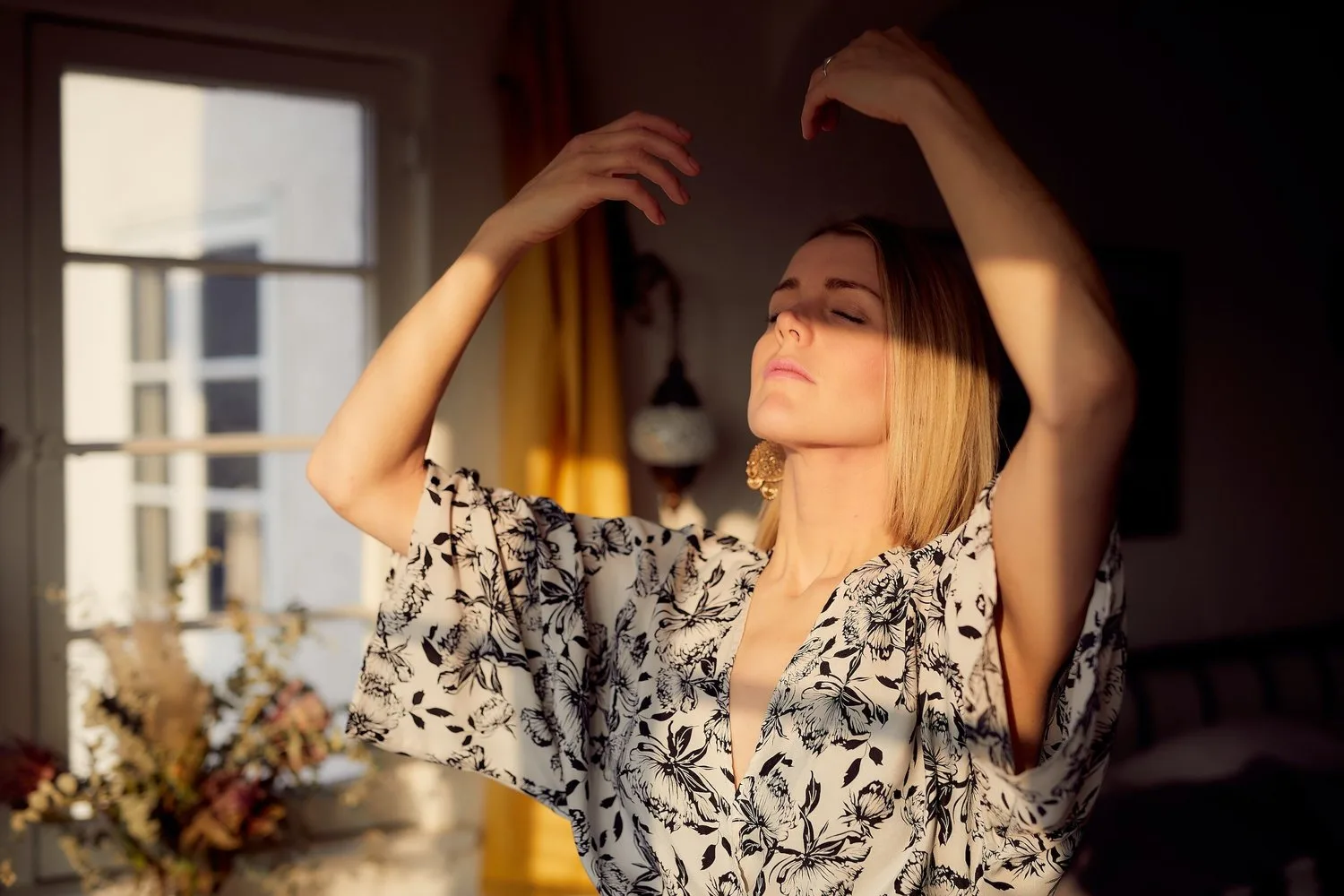 Woman in a floral top raising her arms in warm window light