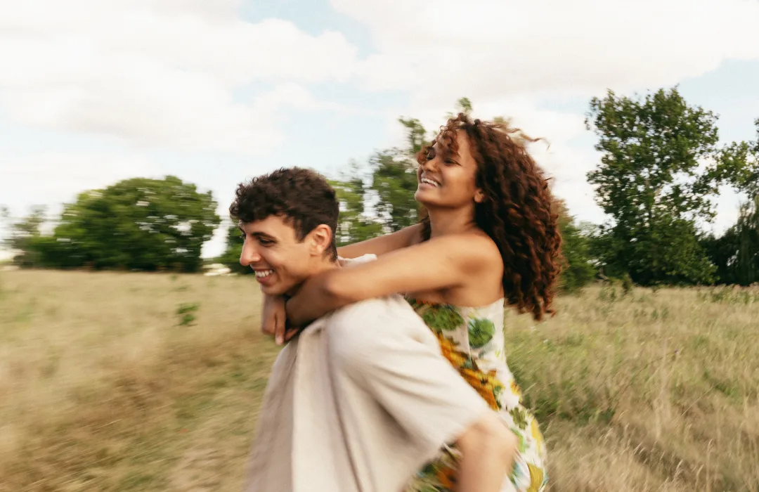 Laughing couple outdoors, one carrying the other piggyback through a field