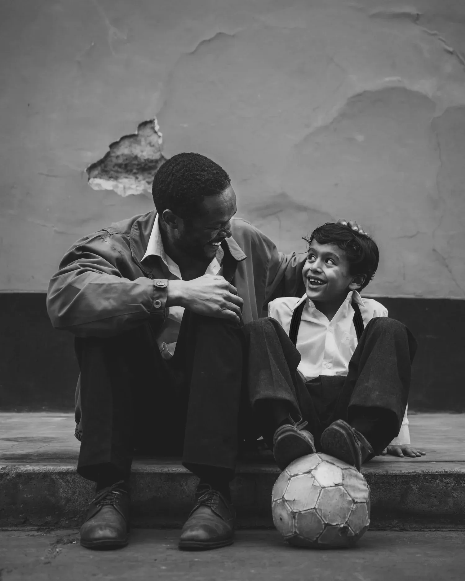 Black-and-white photo of a father and son sitting on a curb with a soccer ball