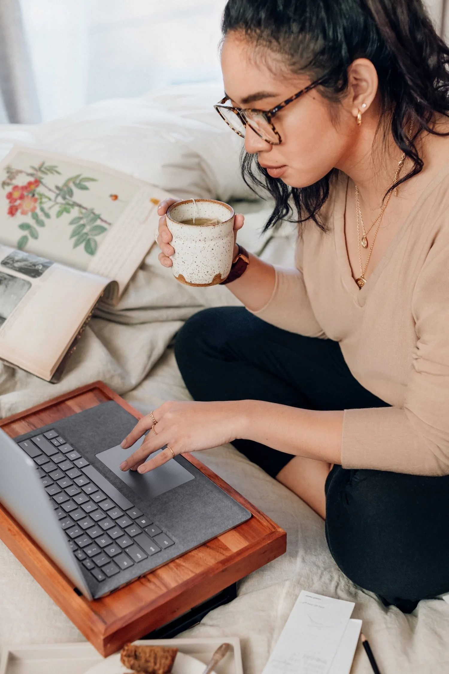 Woman in glasses working on a laptop in bed with a mug beside her