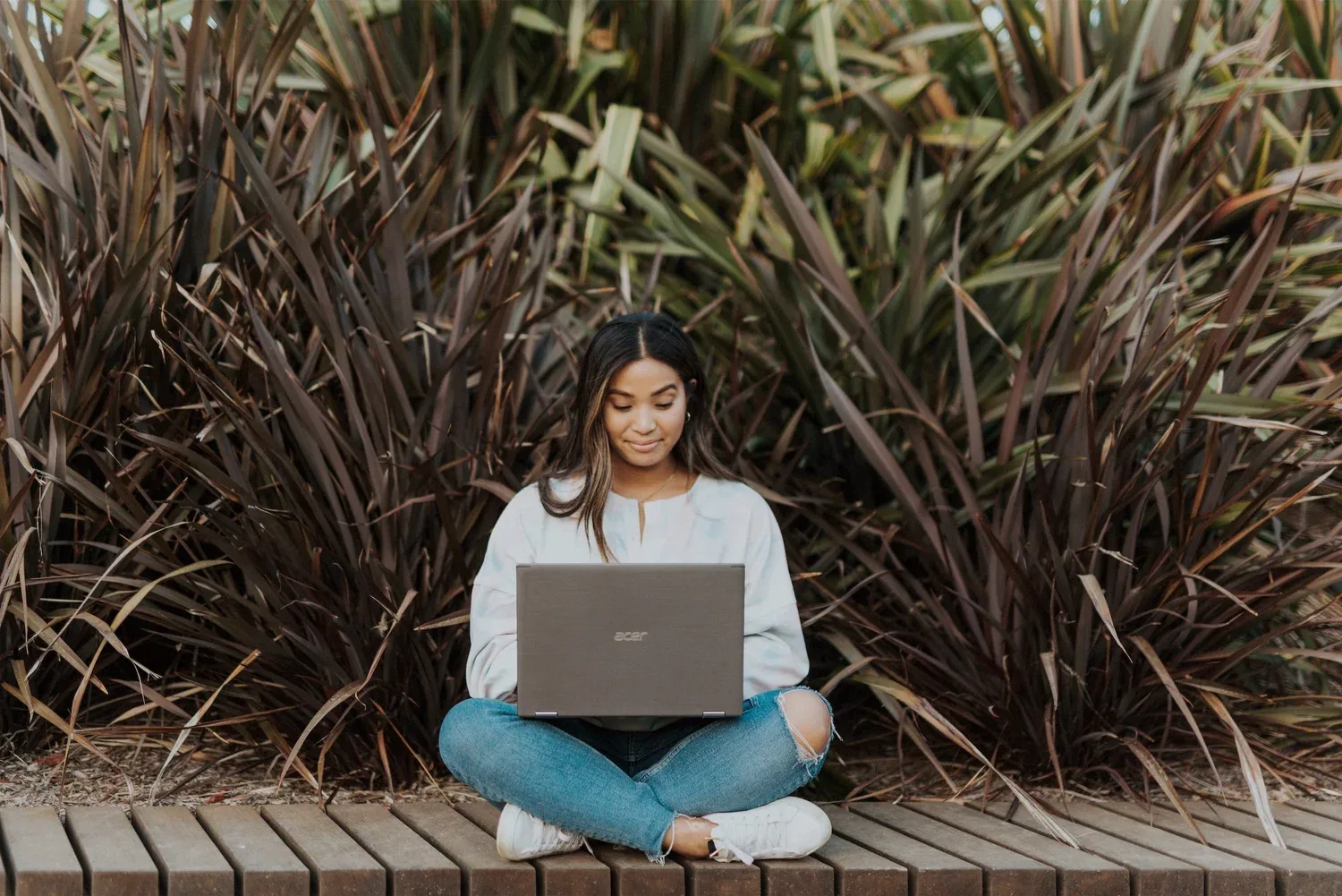 Woman sitting cross-legged outdoors with a laptop in front of tall grasses