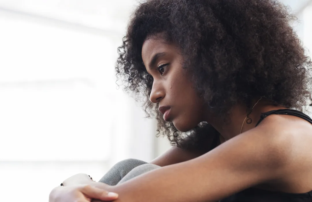 Woman with curly hair sitting on the floor resting her head on her knee, looking pensive