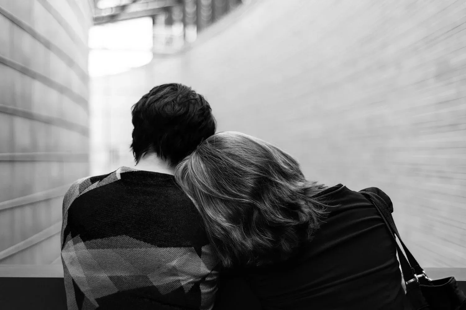 Black-and-white photo of two people sitting close, one resting on the other’s shoulder