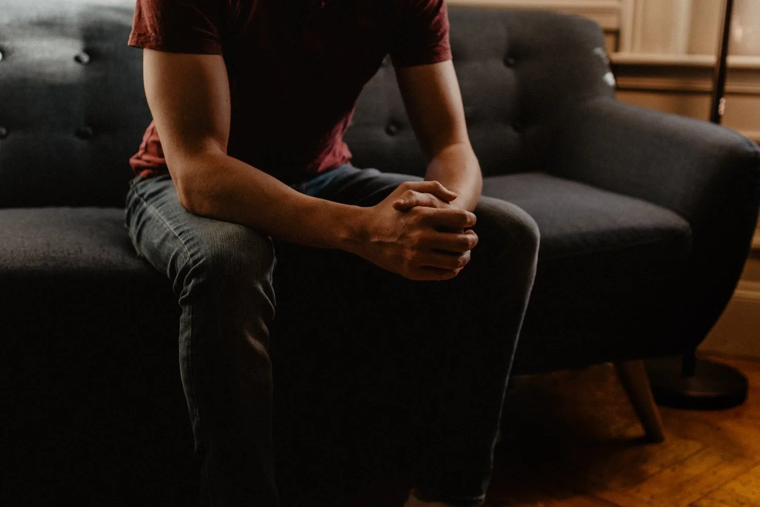 Person seated on a dark sofa with hands clasped between their knees