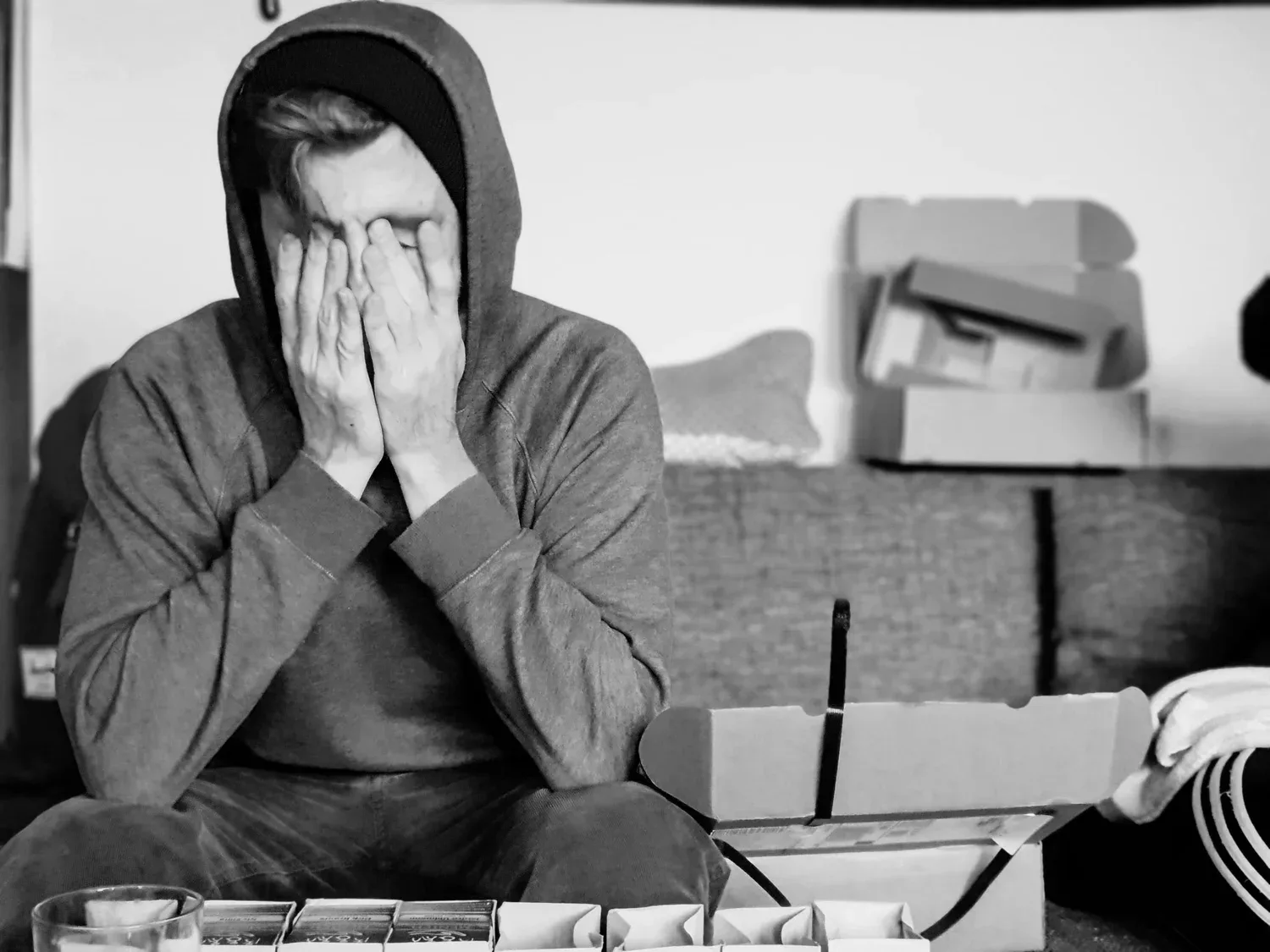 Black-and-white photo of a person in a hoodie covering their face beside moving boxes