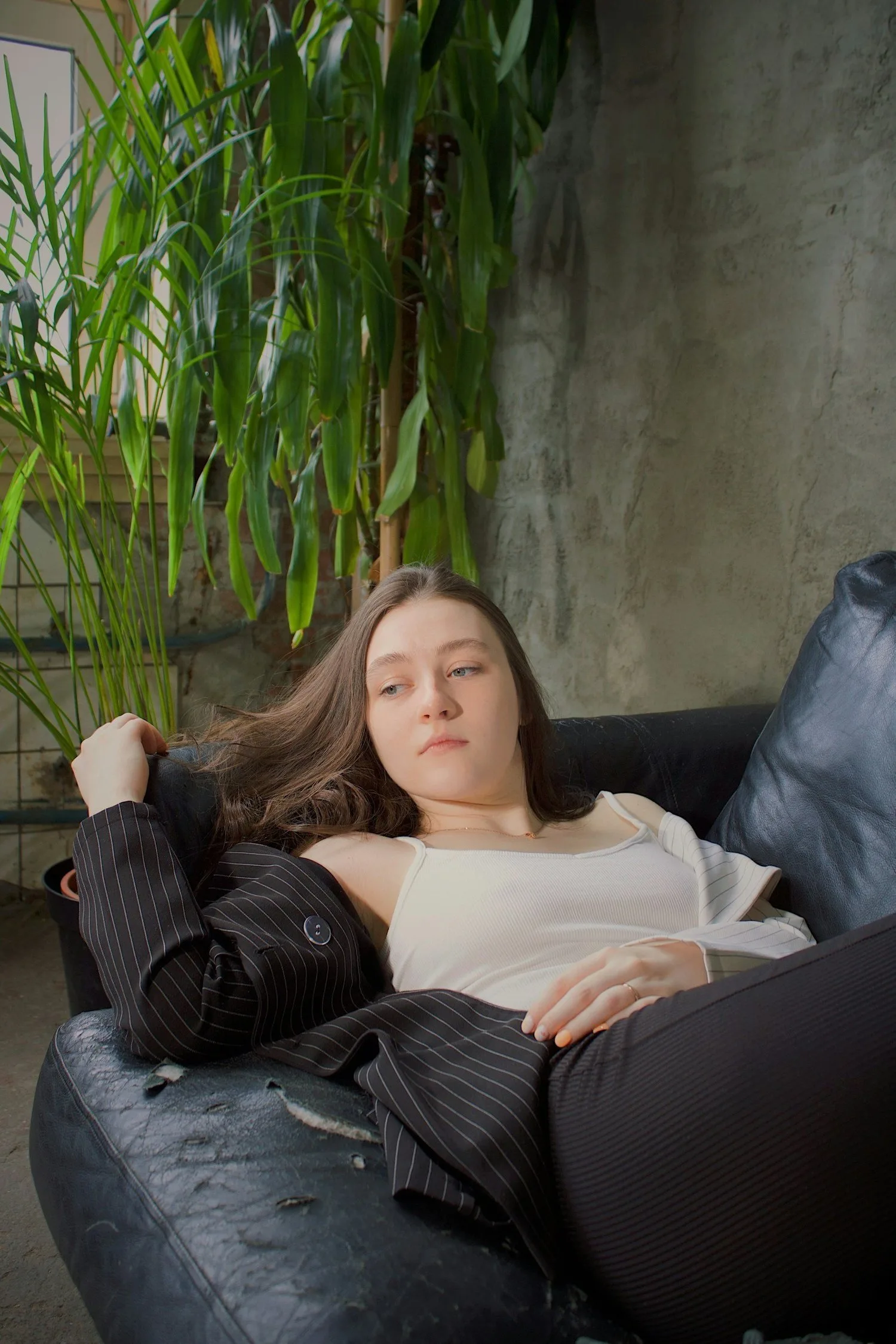 Young woman lying on a dark sofa in a plant-filled room, looking at the ceiling