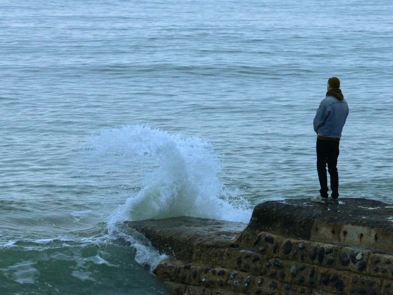 Person standing alone on a rocky shore as a wave crashes nearby