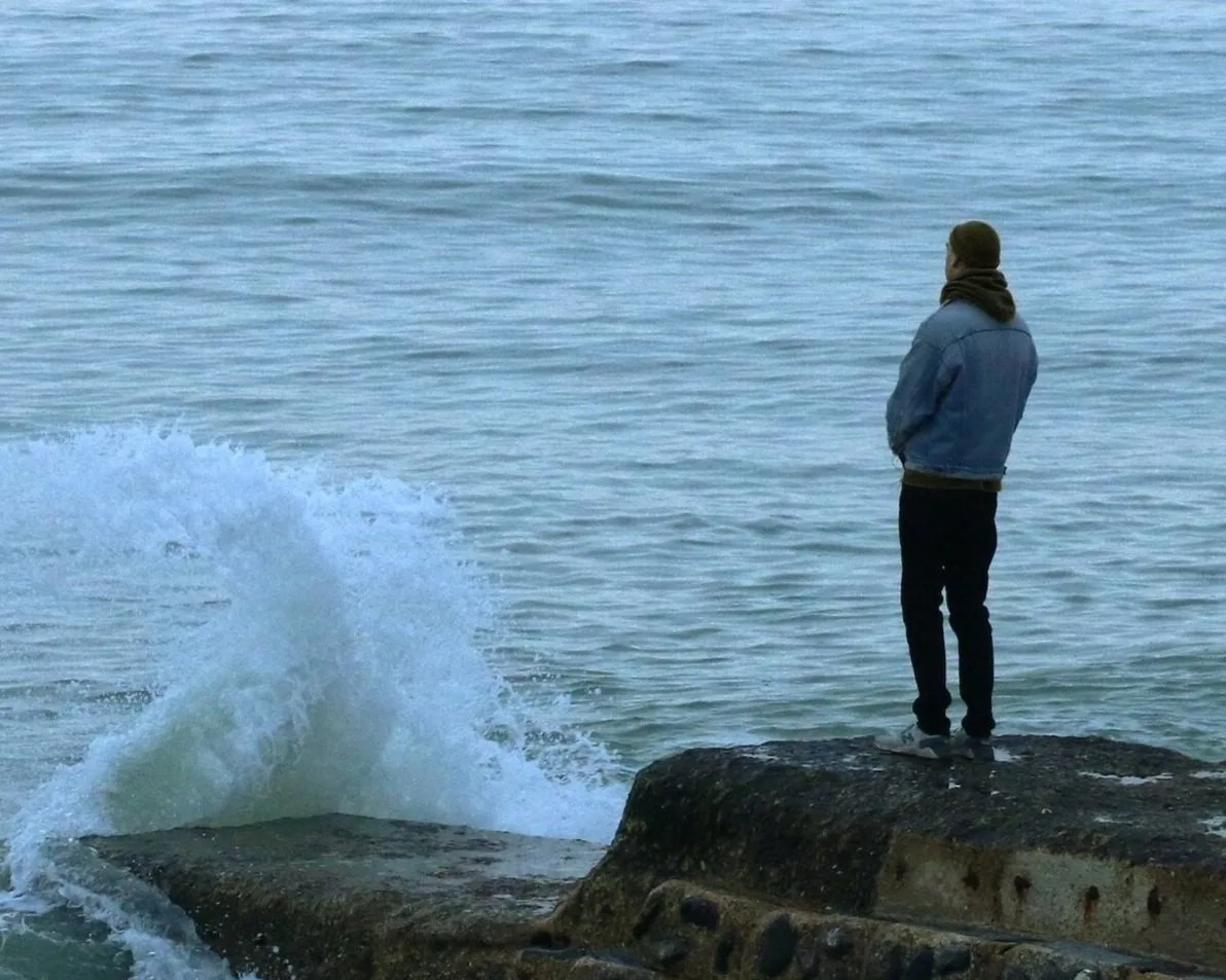 Person standing alone on a rocky shore as a wave crashes nearby