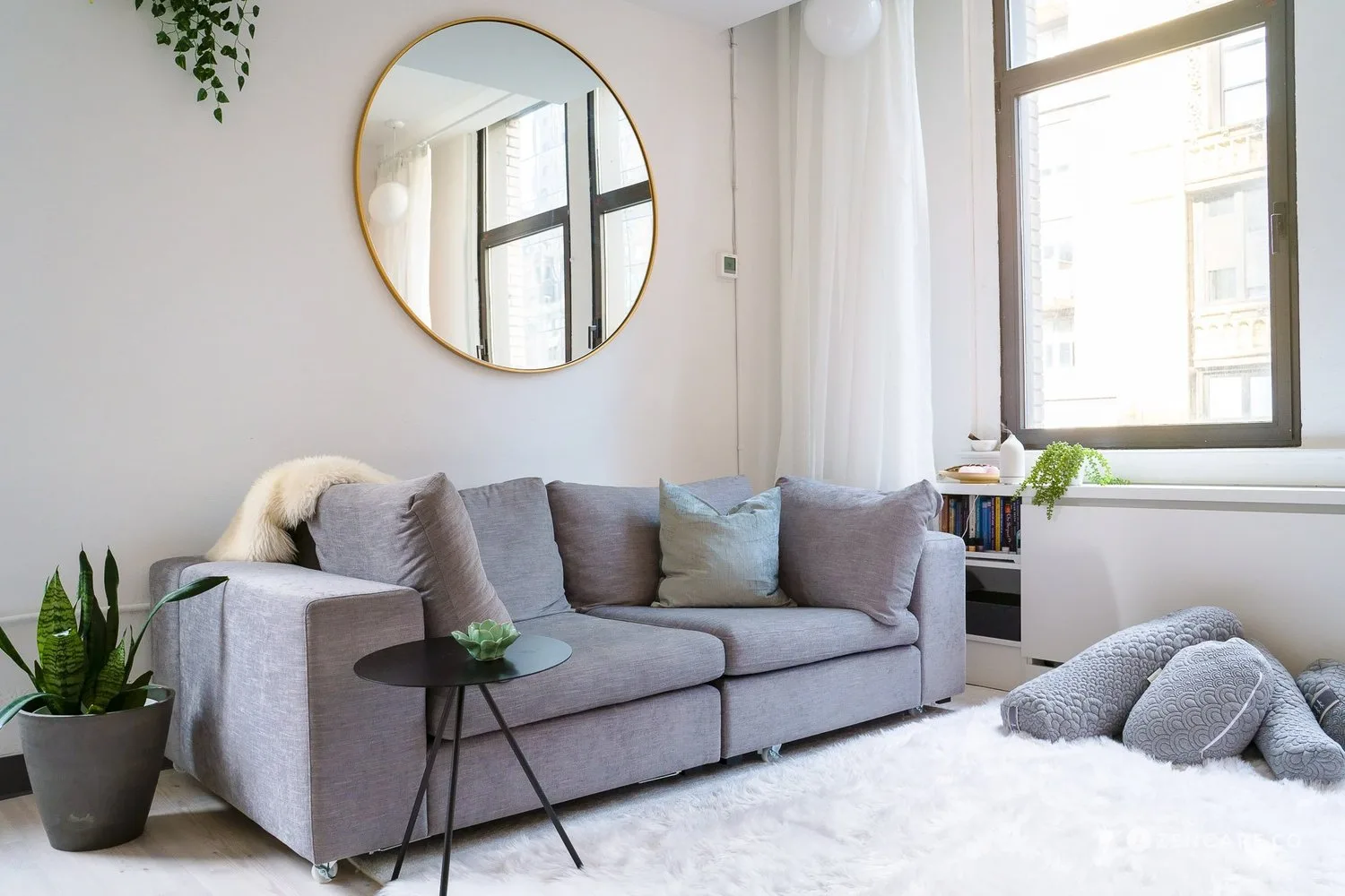 Bright living room with a gray sofa, round mirror, and plants