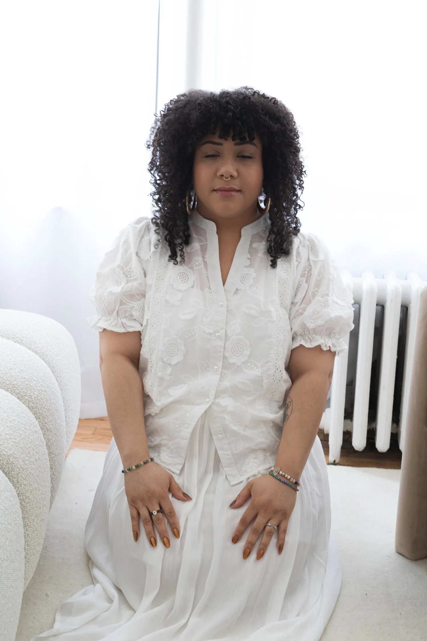 Woman with curly hair in a white dress sitting upright with hands on her knees in a bright room