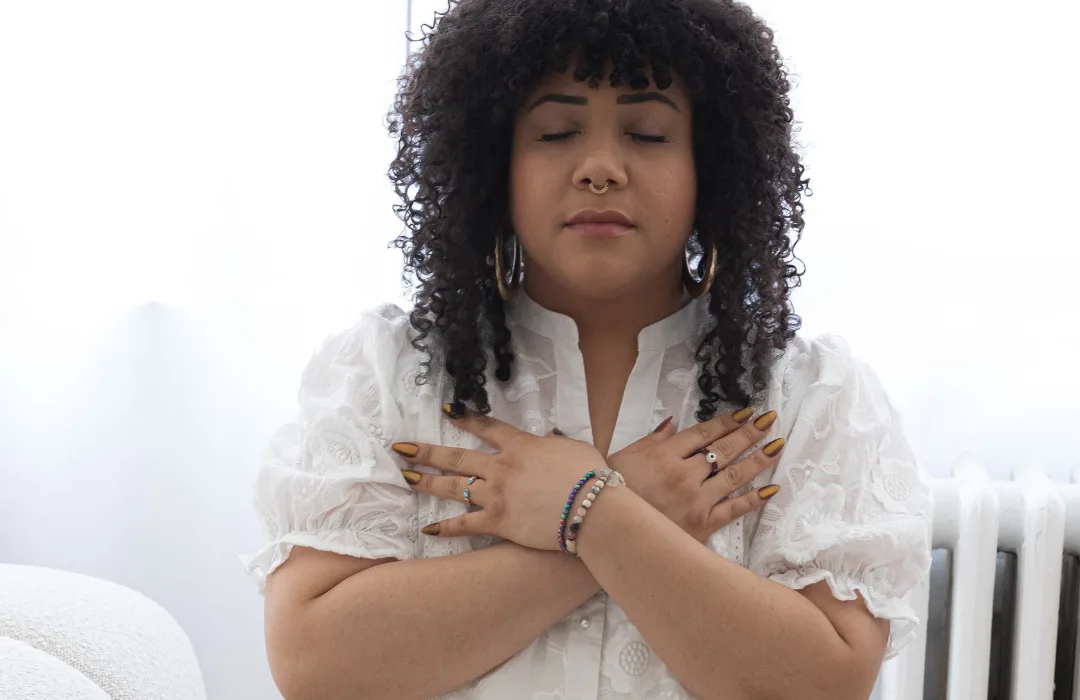 Woman with curly hair in a white dress with arms crossed over her chest in a self-hug