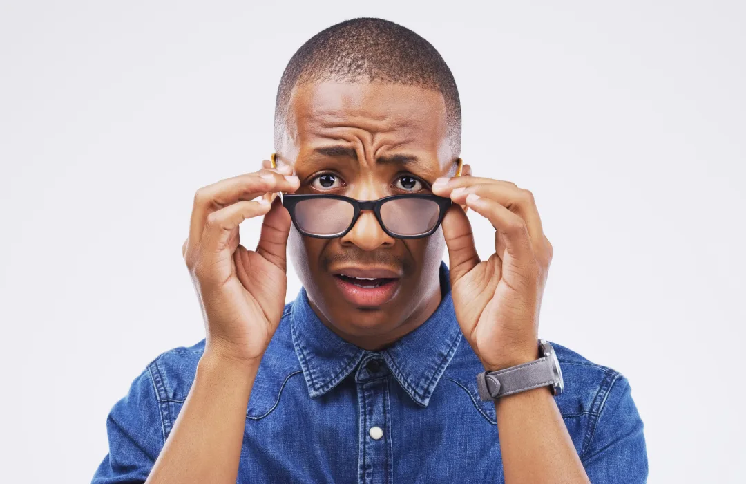 Man in a denim shirt lowering his glasses with a worried, wide-eyed expression