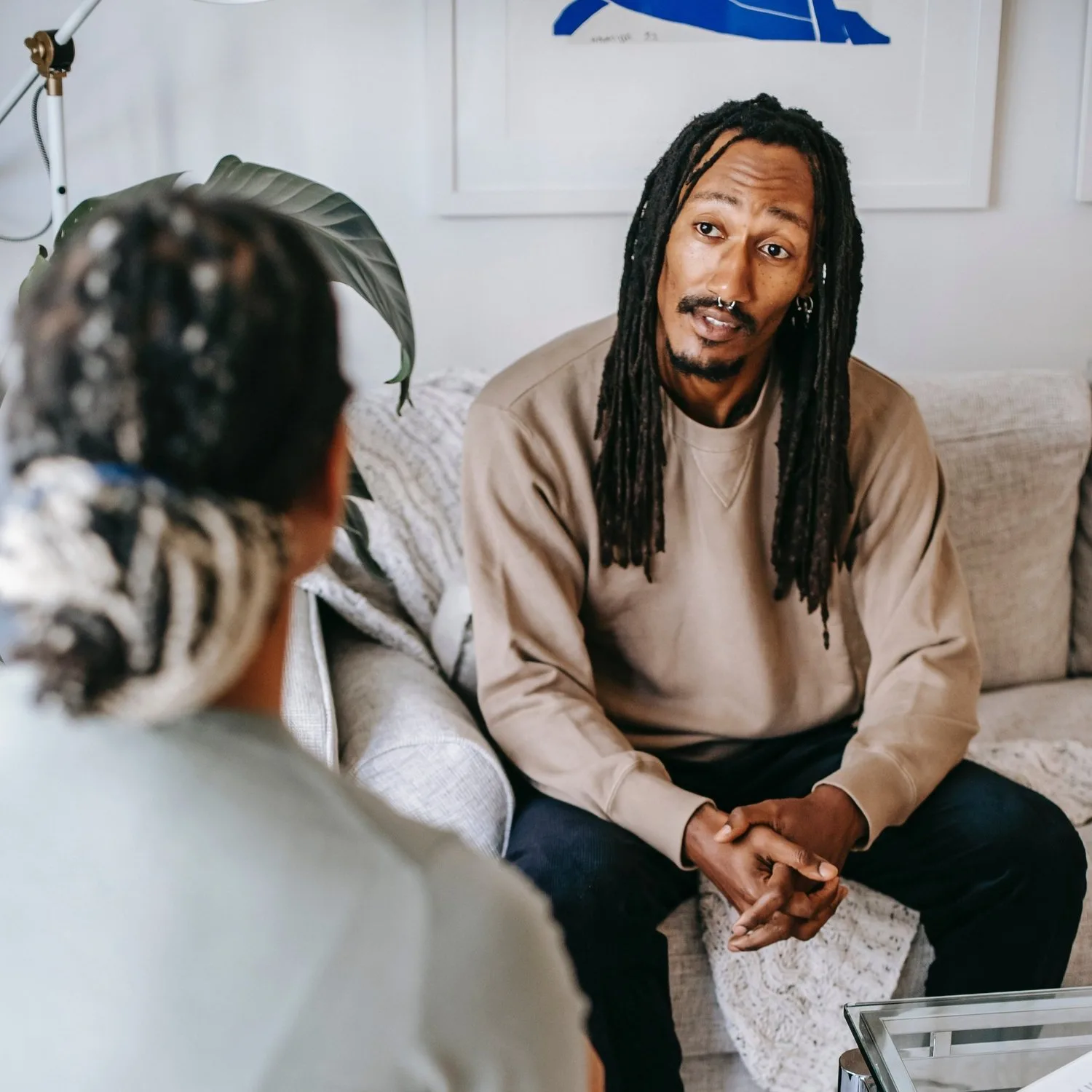 Man with locs seated on a sofa talking with someone across from him