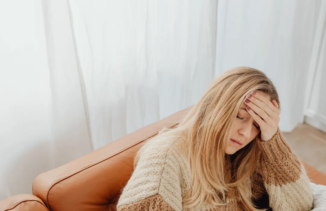 Woman with long hair sitting on a sofa with a hand to her forehead, looking stressed