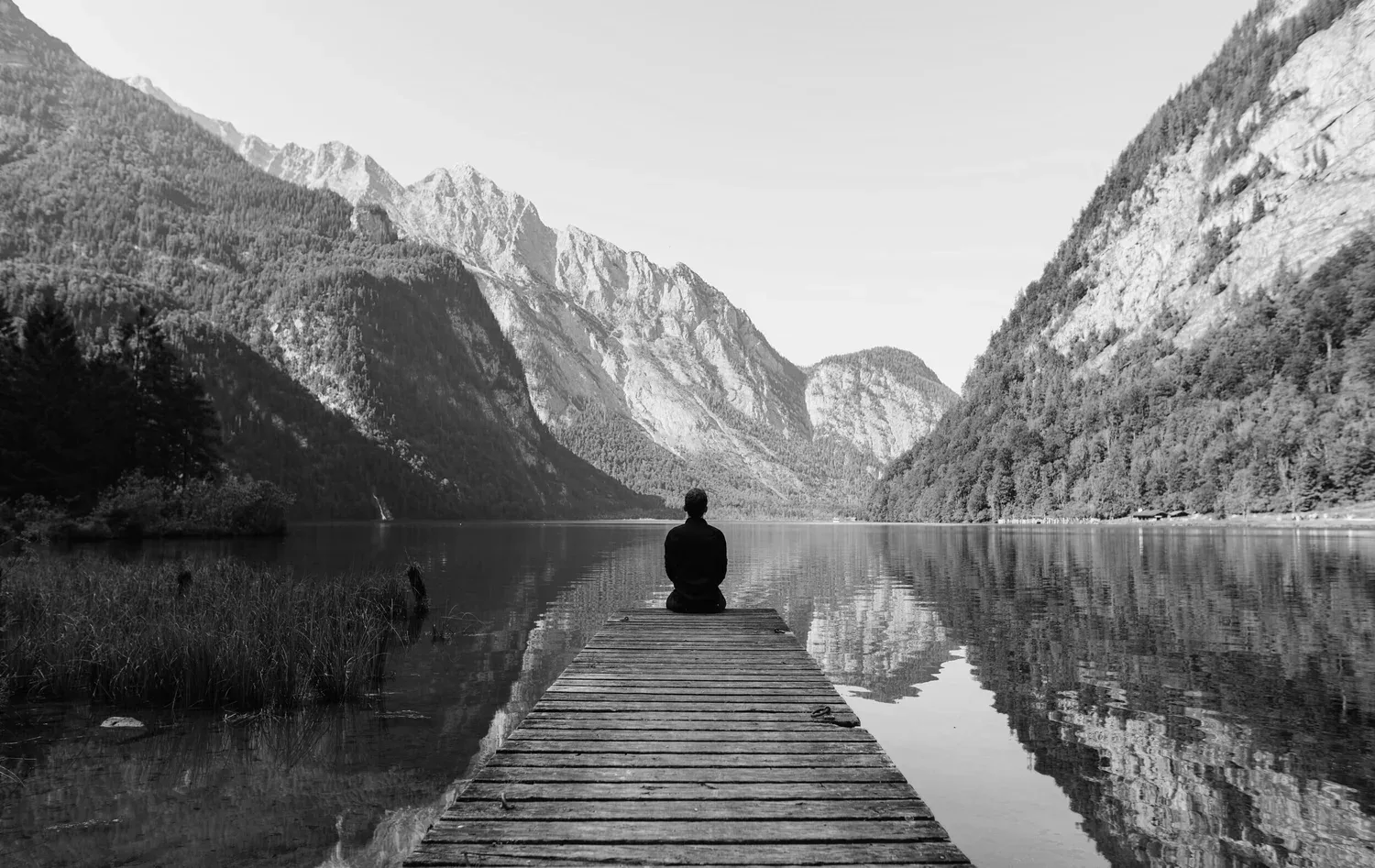 Black-and-white photo of a person sitting at the end of a dock on a mountain lake