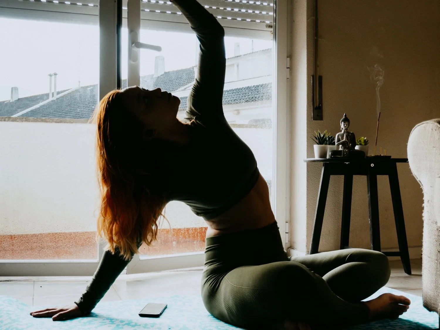 Woman in a seated stretch reaching one arm overhead by a window