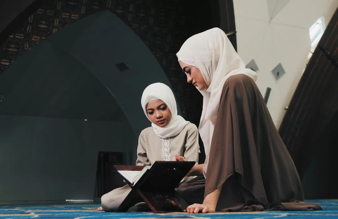 Woman in a hijab reading with a young girl in a hijab seated on a mosque carpet