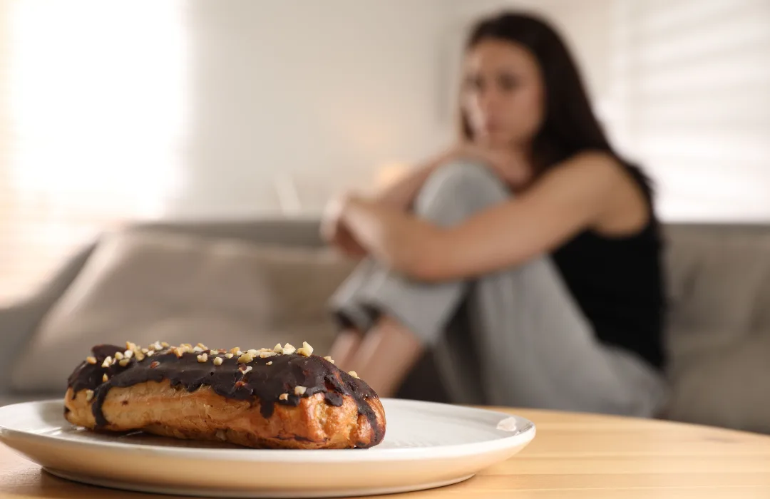 Woman hugging her knees on a sofa with a pastry on a plate in the foreground