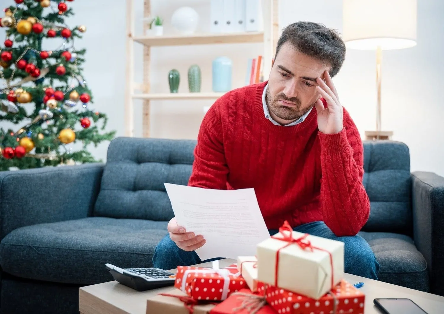 Man in a red sweater frowning at a bill beside a Christmas tree and gifts