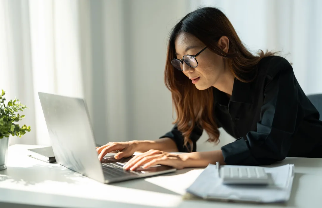 Woman in glasses working intently at a laptop in a bright home office