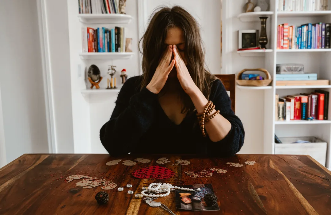 Woman at a wooden table with hands pressed together near her face, bookshelves behind her