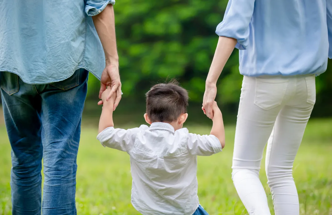 Child holding hands with two adults while walking across a grassy lawn