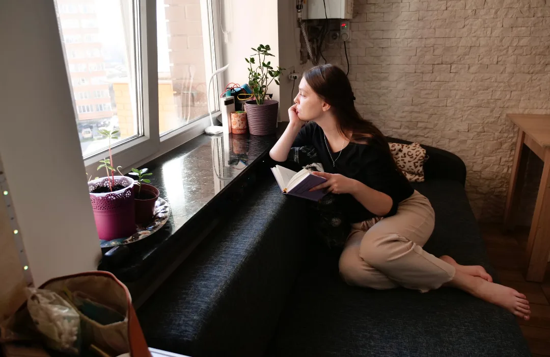 Woman sitting on the floor by a window, looking out thoughtfully