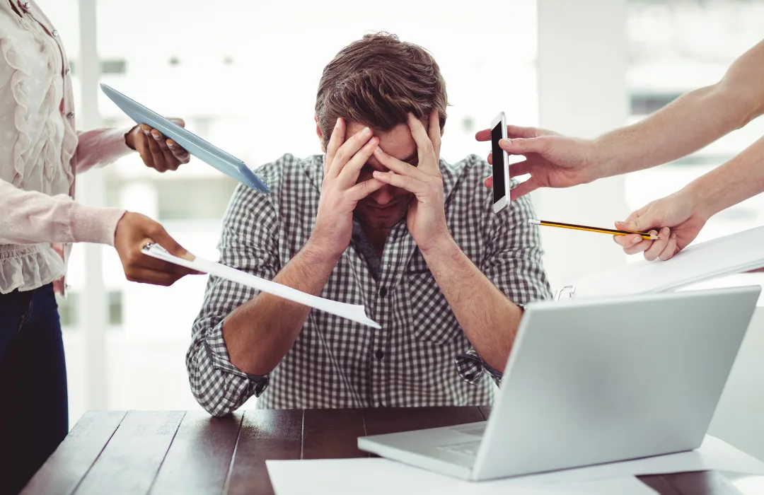 Overwhelmed man at a laptop with his head in his hands while papers and phones are pushed at him