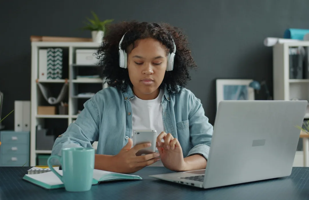 Young woman with headphones using her phone at a desk with a laptop
