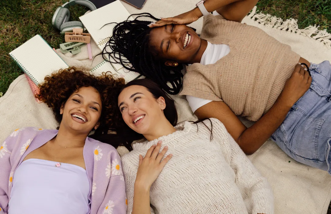 Three young women lying on a blanket with books, smiling up at the camera