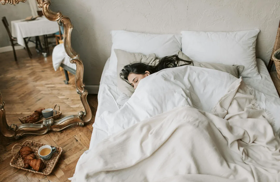 Person curled up asleep under white bedding in a softly lit bedroom