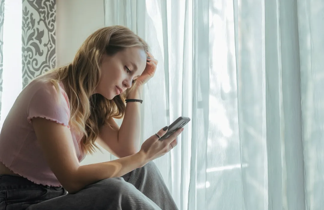 Woman sitting by a window looking down at her phone with a pensive expression