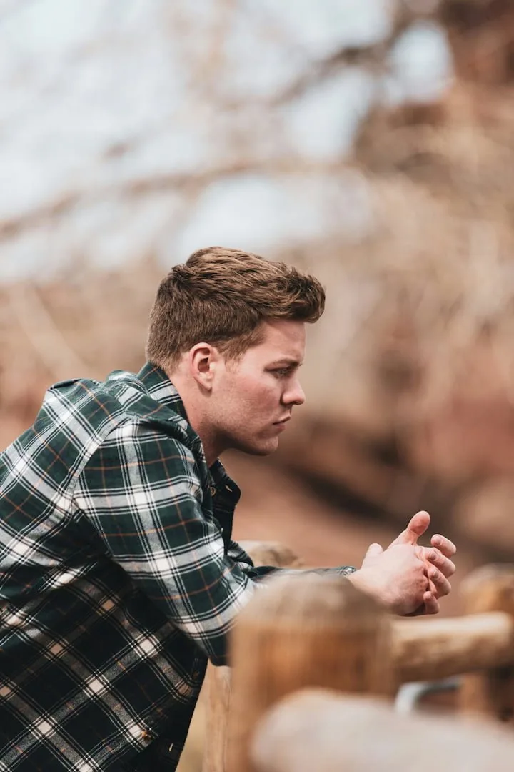 Man in a flannel shirt sitting outdoors, warming his hands and looking into the distance