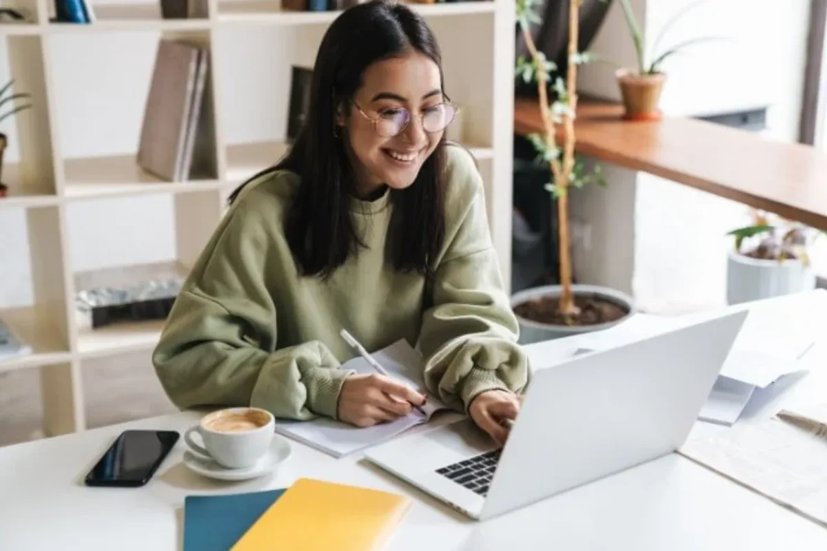 A smiling teen writing notes beside a laptop at a bright desk